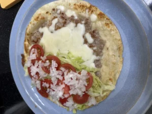 또띠아 위에 구운 고기 패티, 녹은 모짜렐라 치즈, 잘게 썬 양상추, 토마토, 다진 양파가 올라가 있는 접기 직전의 모습