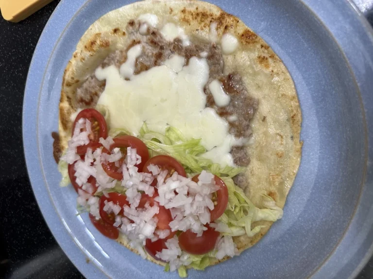 또띠아 위에 구운 고기 패티, 녹은 모짜렐라 치즈, 잘게 썬 양상추, 토마토, 다진 양파가 올라가 있는 접기 직전의 모습
